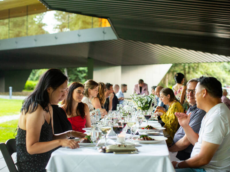 Guests enjoying a summer long-table dinner beneath the Audain Art Museum in Whistler, with elegant décor, wine glasses, and vibrant conversation in a unique architectural setting.