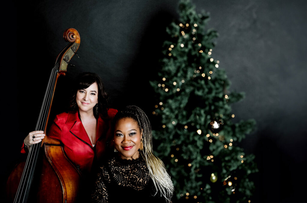 Two award-winning powerhouses, vocalist Dawn Pemberton and bassist/bandleader Jodi Proznick pose in front of a Christmas Tree.