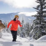 Two people snowshoe in thick snow with Whistler Blackcomb in the background. They smile as the sun comes out.