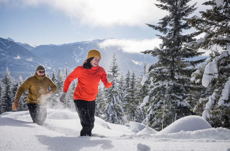 Two people snowshoe in thick snow with Whistler Blackcomb in the background. They smile as the sun comes out.