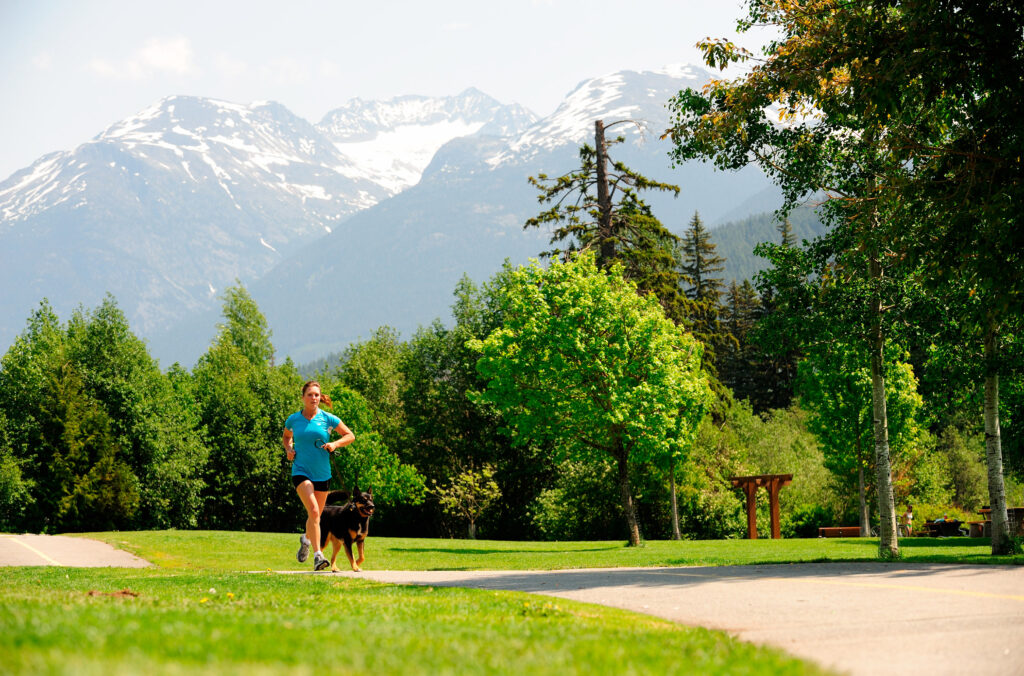 A person runs with their leashed dog along the Whistler Valley Trail with the mountains in the distance.