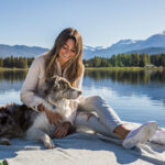 A woman sits with her dog on the shores of a lake in Whistler.