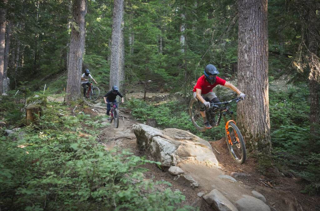 Three downhill mountain bikers tackle some of the blue and black technical features in the Creekside Zone of the Whistler Mountain Bike Park.