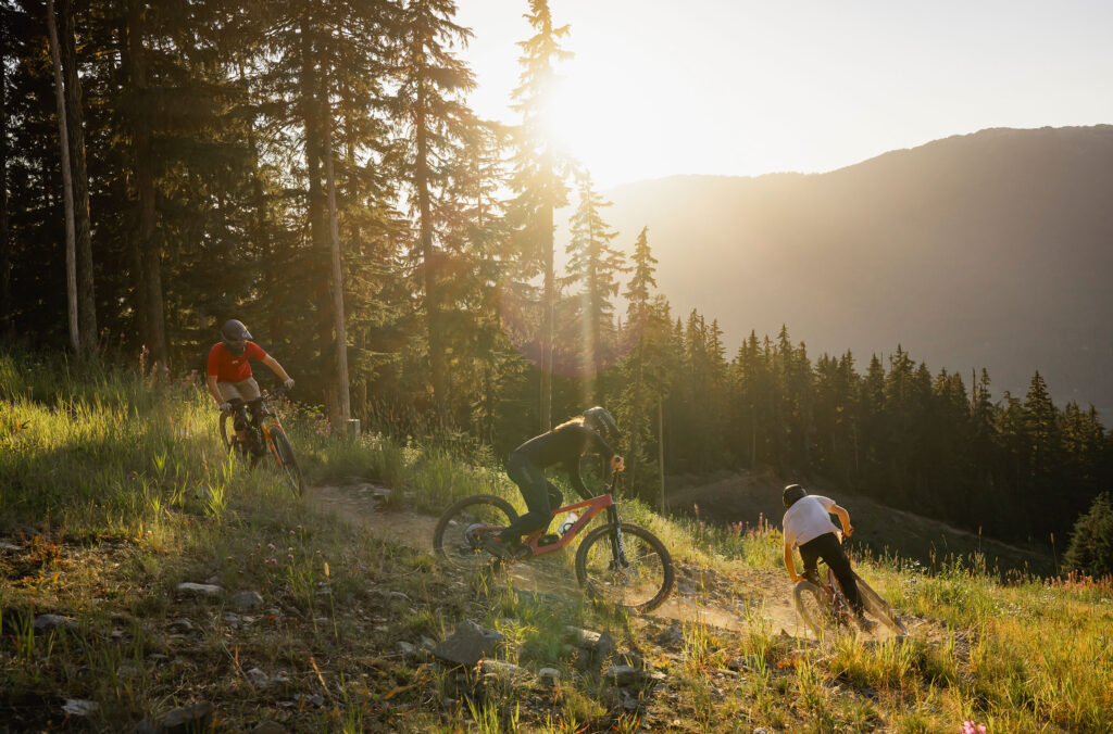 Three downhill mountain bikers enjoy the Creekside Zone of the Whistler Mountain Bike Park as the summer sun sets.