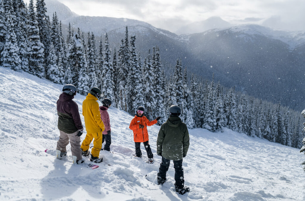 A snowboard instructor explains to a group of snowboarders what they're going to tackle next.