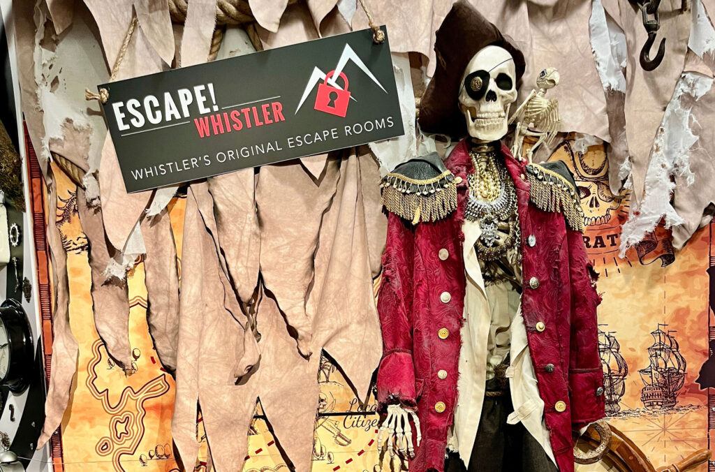 The photo backdrop for the Buried Treasure escape room has a skeleton dressed in pirate gear with a wooden chest at its feet.