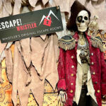 The photo backdrop for the Buried Treasure escape room has a skeleton dressed in pirate gear with a wooden chest at its feet.