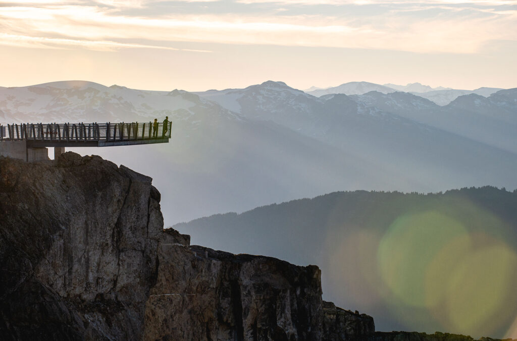 Two people stand on the Raven Eye on Whistler Blackcomb, looking out over the Coast Mountains in the summer sun.