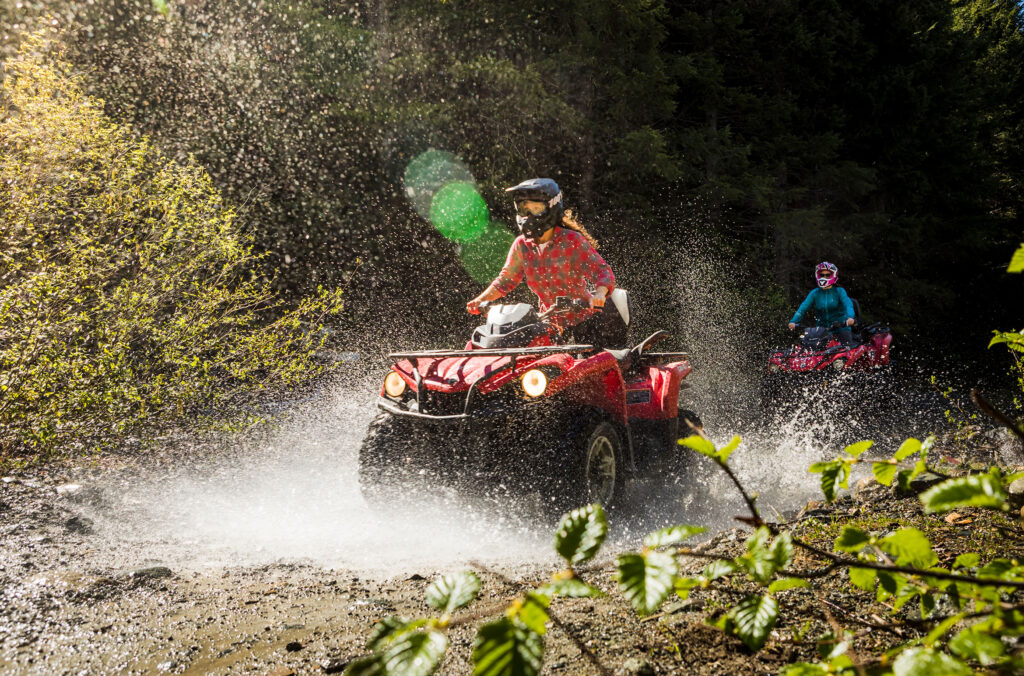 A bright red ATV splashes through a puddle, water flying. The rider is wearng a full face helmet with a plaid shirt, her hair is flying.