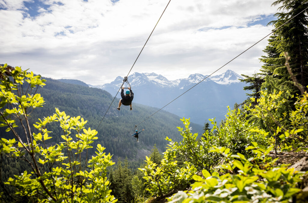 Two zipliners whizz across a lush, green valley on a double zipline.