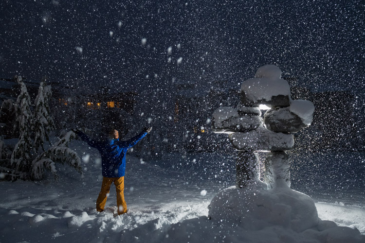 A man in snow gear, stood next to a snow-laden Inuksuk raises his arms to the evening sky as the snow falls in Whistler.