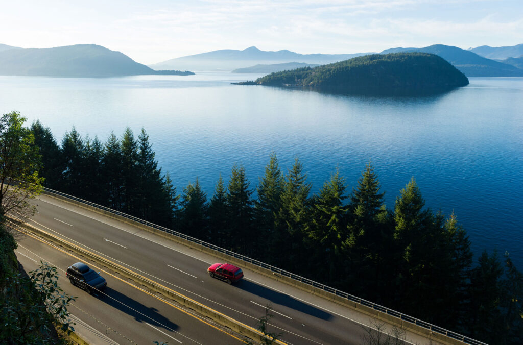 A shot of the Sea to Sky Highway in the summer, with the mountains on the left and ocean on the right.