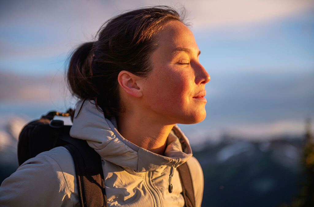 A hiker faces the sun and closes her eyes at the top of the mountain.