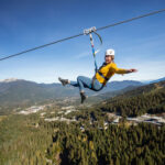 A person ziplines across the valley in between Whistler and Blackcomb mountains, the longest zipline in NA. Arms out, big smile, summer sun, it looks thrilling.