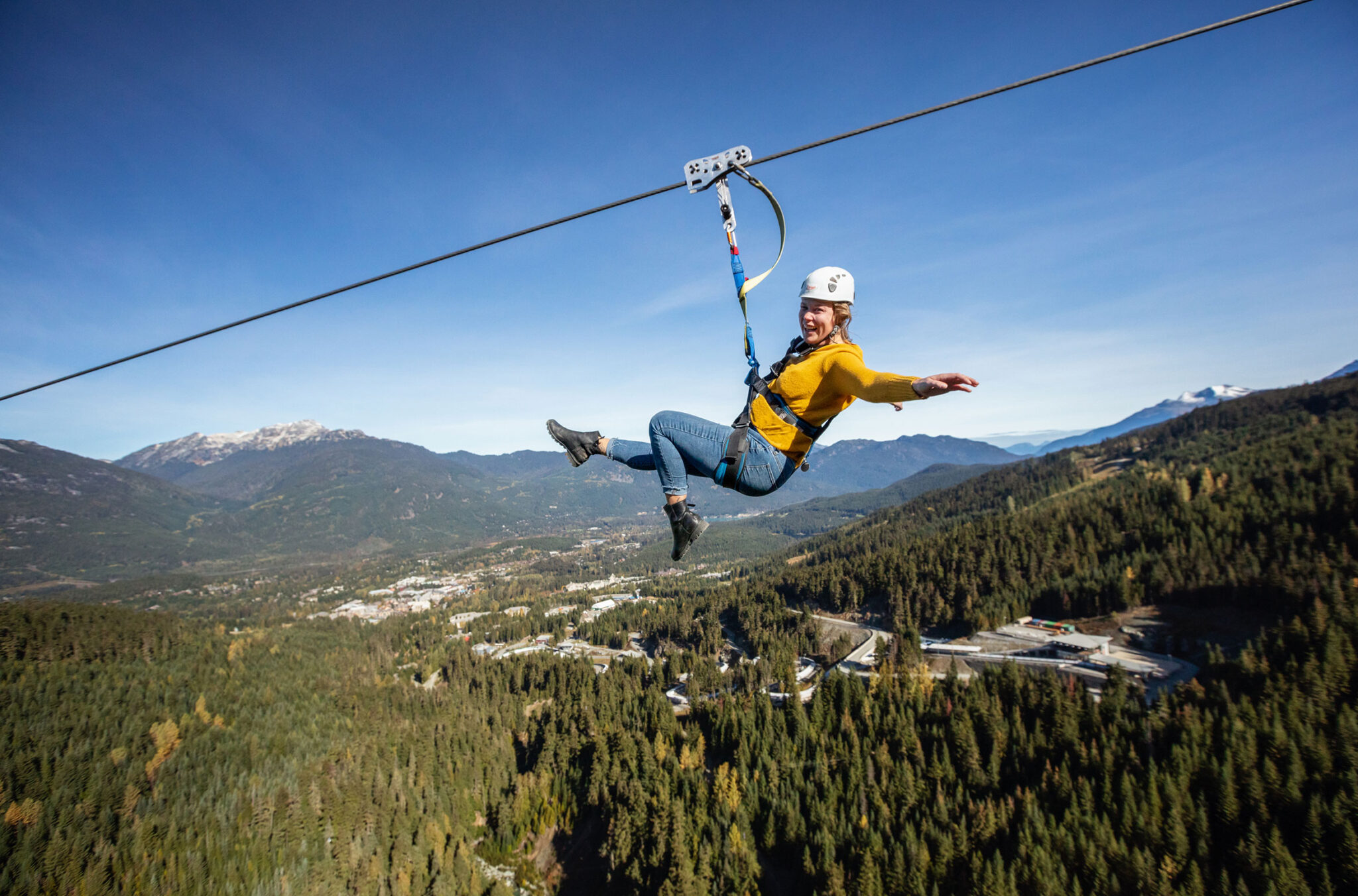 A person ziplines across the valley in between Whistler and Blackcomb mountains, the longest zipline in NA. Arms out, big smile, summer sun, it looks thrilling.
