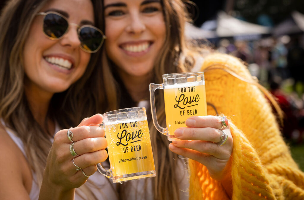 Two women holding beer sample mugs at the Whistler Village Beer Festival, smile at the camera.