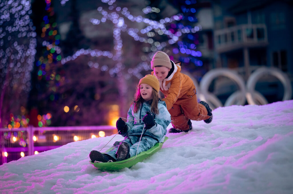 Two kids play with sleds at the Snow Zone at Whistler Olympic Plaza. One is pushing the other down an snowy slope.