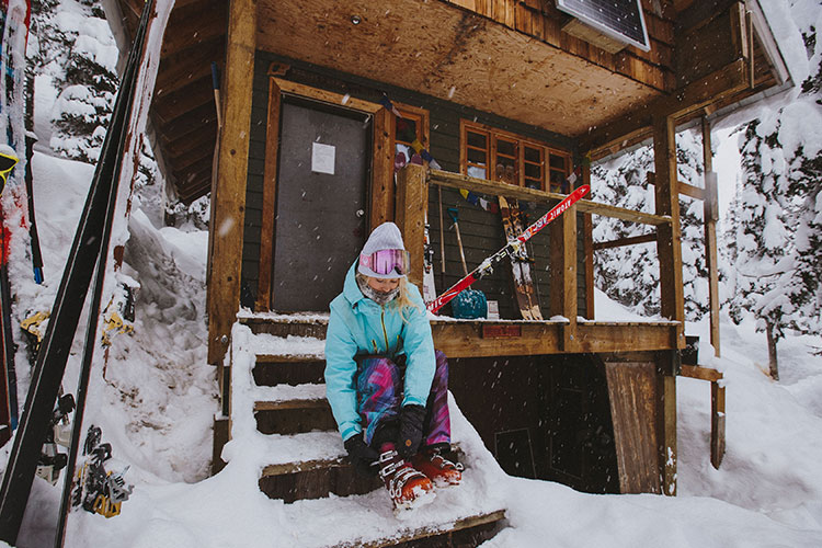 A female skier does up her boots on the steps of a backcountry hut.