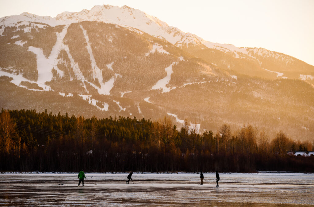 Hockey players skate on a frozen lake in front of a snowy ski hill in Whistler.
