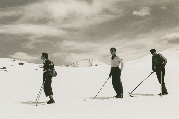 Black and white image of three male skiers in the mountains in sweaters, socks, and sunglasses.