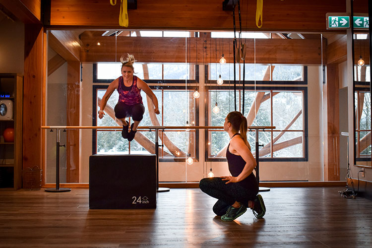 Nina jumps off a box in a gym while her trainer encourages her.