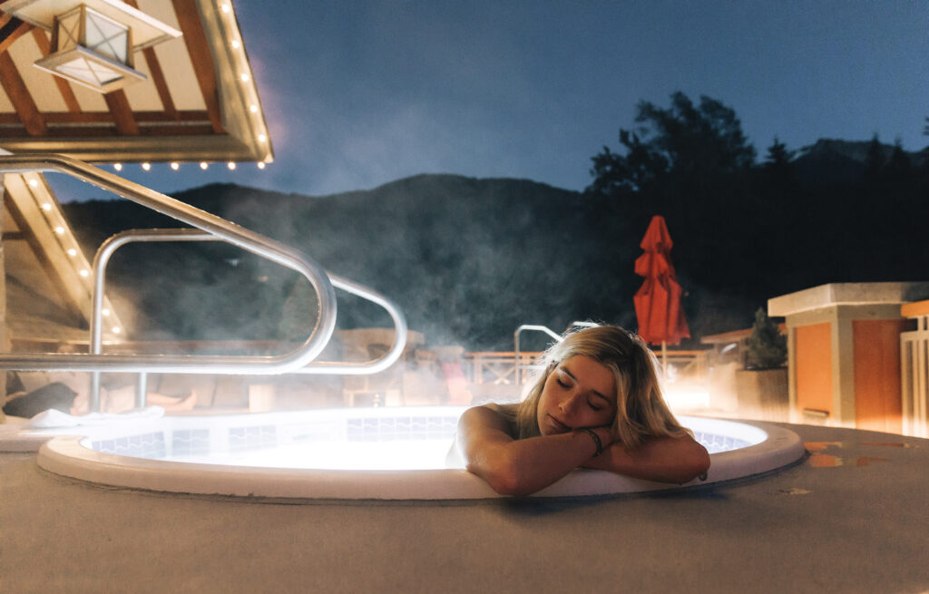 A person relaxes in an outdoor hot tub at night resting their arms on the edge of the tub and closing their eyes.