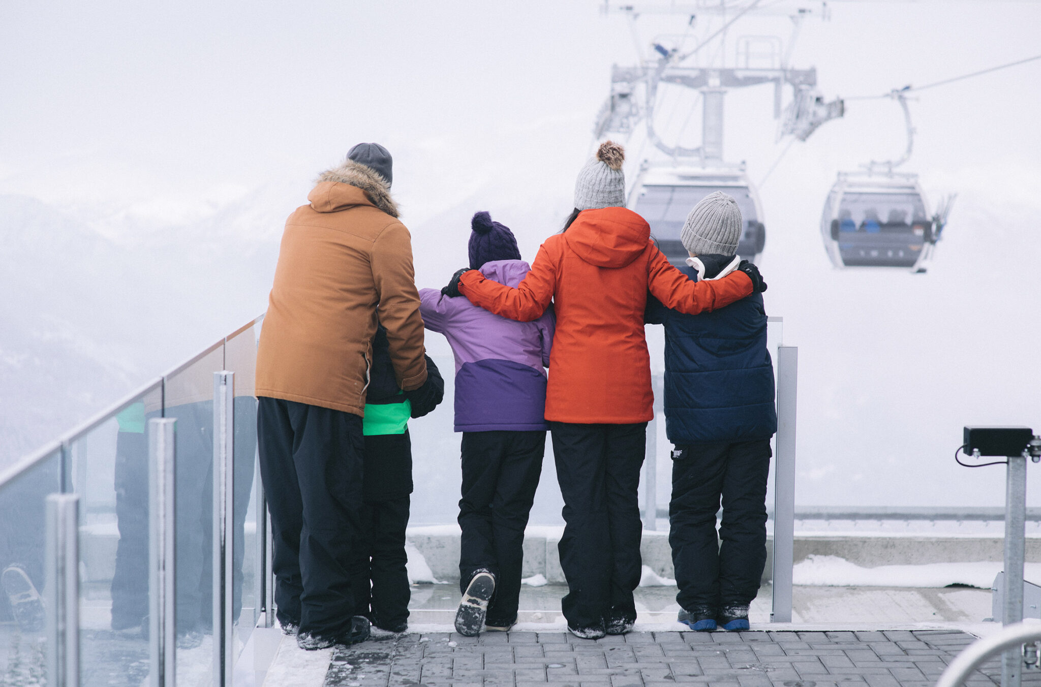 A family sightseeing in winter on Whistler Blackcomb stand looking out at the gondolas coming in.