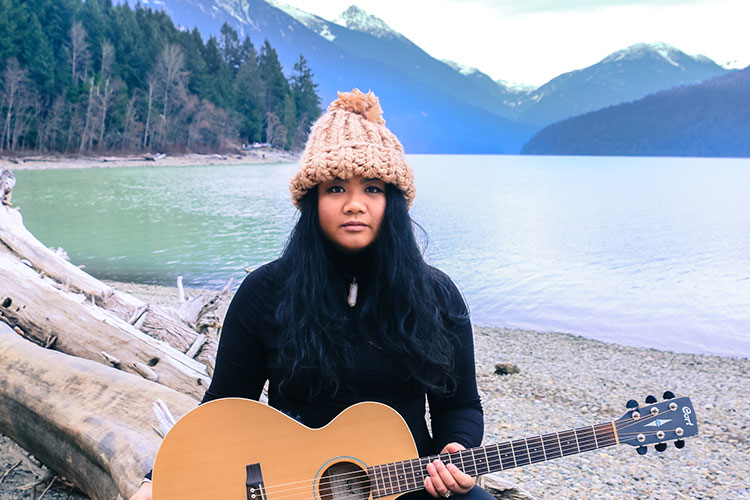 Musician Jenna Mae looks at the camera holding her guitar with a Whistler Mountain backdrop.