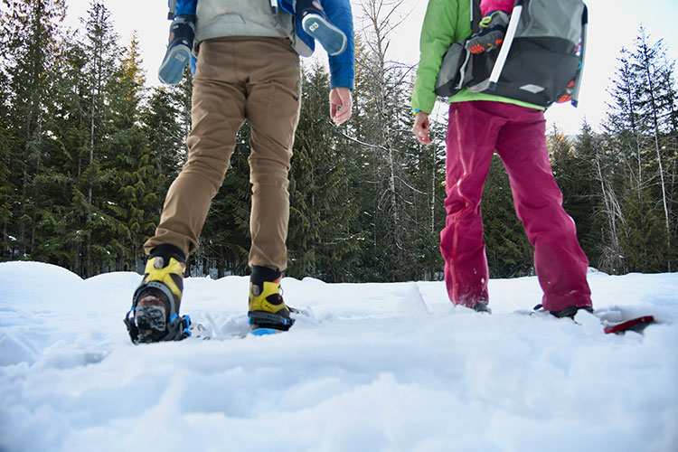 Four sets of legs, two dangle from backpacks. Parents are taking their children on a snowshoe adventure in Whistler.