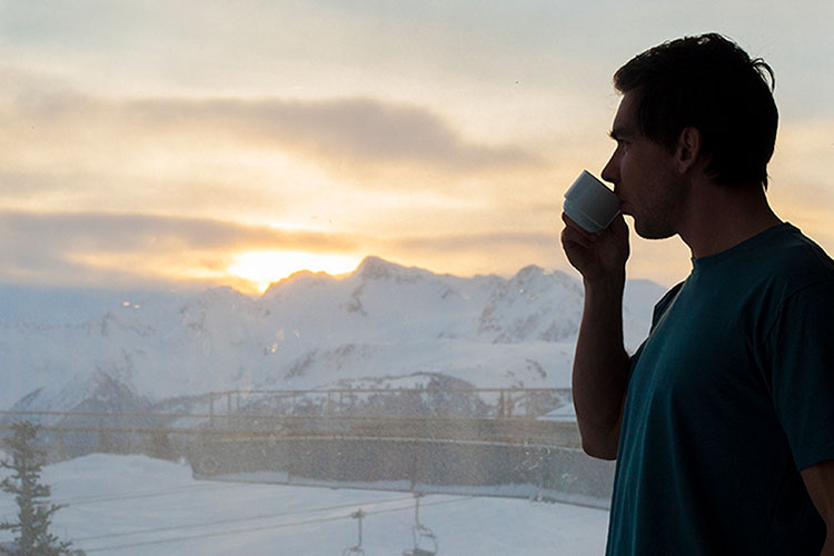 A man drinks coffee at the Roundhouse Lodge on Whistler Mountain overlooking the mountains at sunrise.