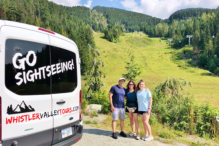 A family pose for a photo while on a sightseeing tour in Whistler with the Olympic rings and ski hill in the background.