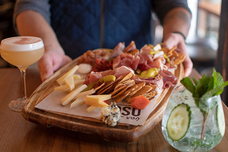 A man holds a charcuterie board filled with meats, cheese, chutneys and crackers in between two cocktails at Bar Oso in Whistler.