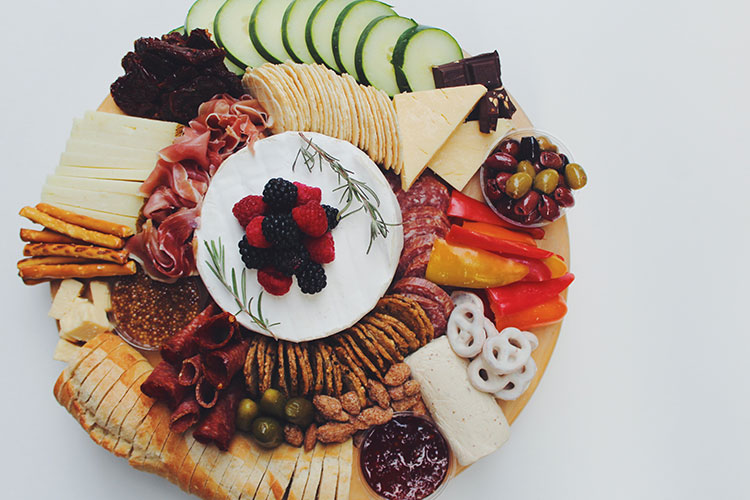 A round charcuterie platter with cheeses, crackers, veggies, olives, meats and sweet treats.