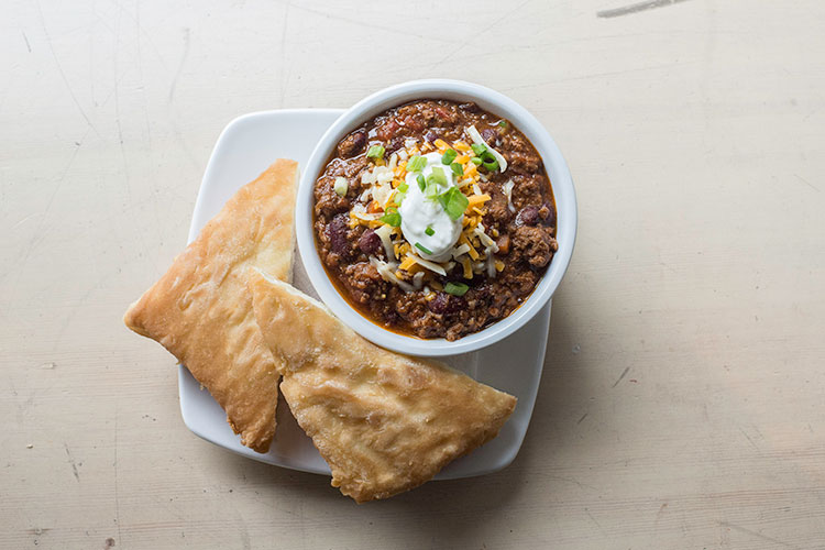 A bowl of chilli topped with sour cream, cheese and chives with a side of bannock bread.