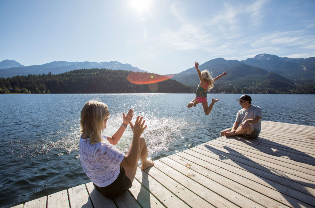 A young girl jumps into a Whistler lake in the sunshine as her parents cheer her on.