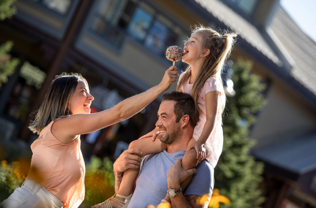A mother feeds her little girl an ice cream while she sits on her dad's shoulders on the Whistler Village Stroll.