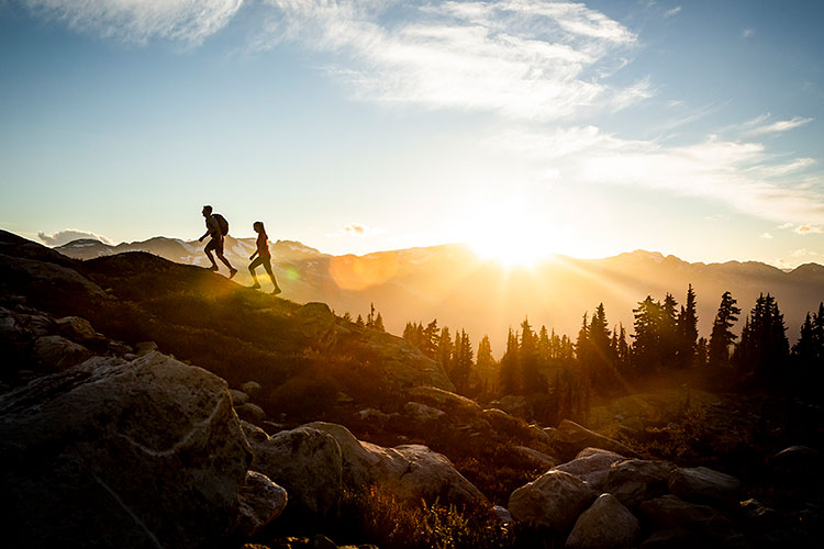 Two people hike on Whistler Blackcomb in the high alpine. The sun rises in the background.