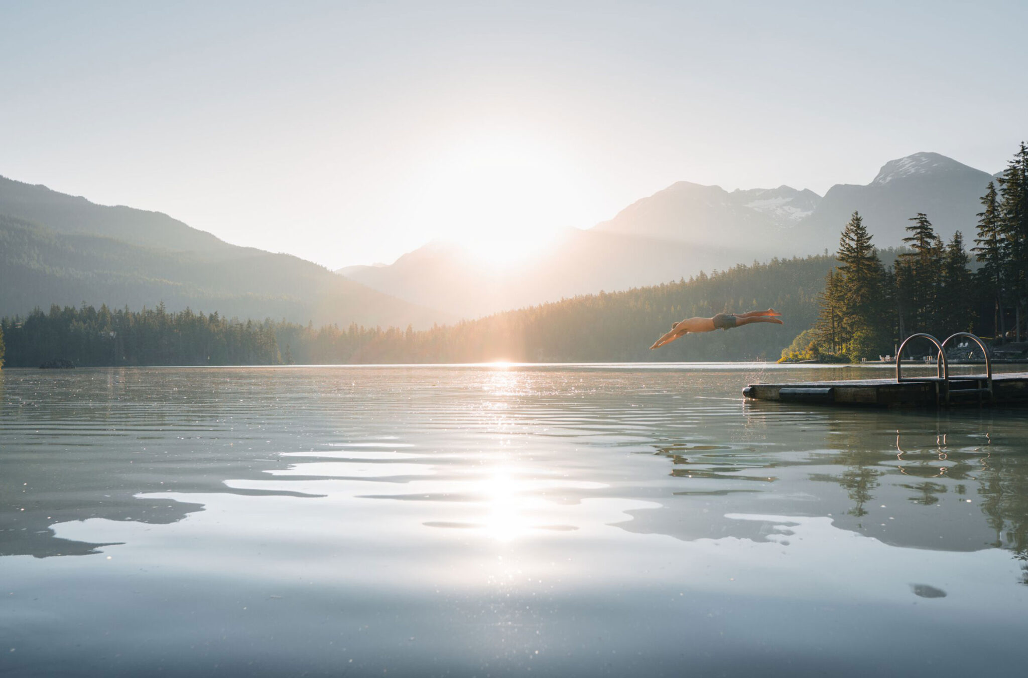 A person dives off the edge of a dock into the mirror-like waters of a Whistler lake in the summer sun.