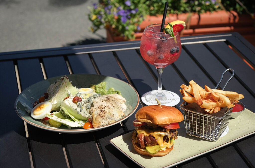 The Feeling Peachy cocktail sits on a sunny patio table flanked by a generous cobb salad and stacked burger. 