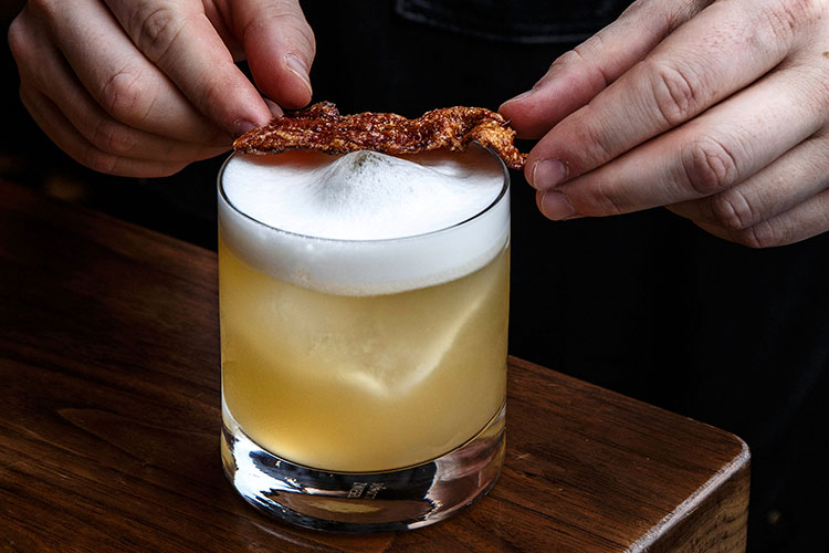 A bartender places a piece of maple bacon on top of the Bar Oso Sour cocktail.