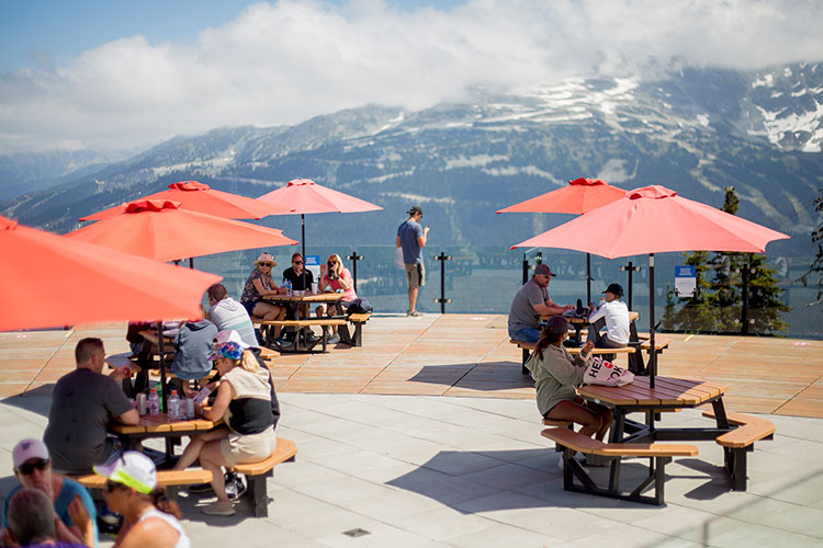 Picnic tables with red umbrellas on a large patio with mountain views on Whistler Blackcomb.