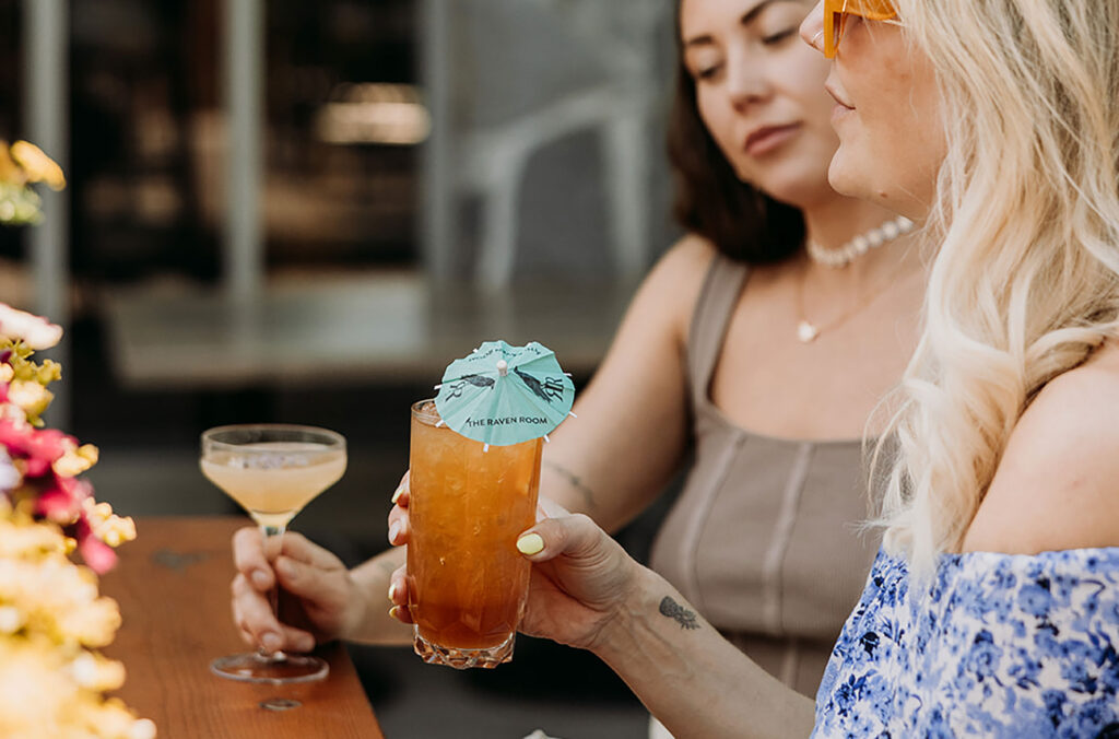 Two women enjoy their summer cocktails on The Raven Room patio overlooking Whistler and Blackcomb mountains.