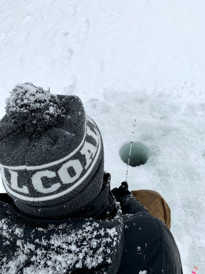 A person sits above a small hole in the ice, fishing with a rod.