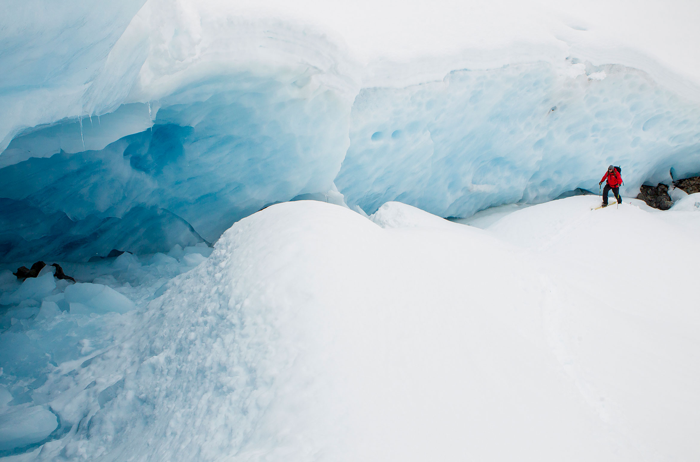 A skier tourer walks by a blue-hued ice wall.