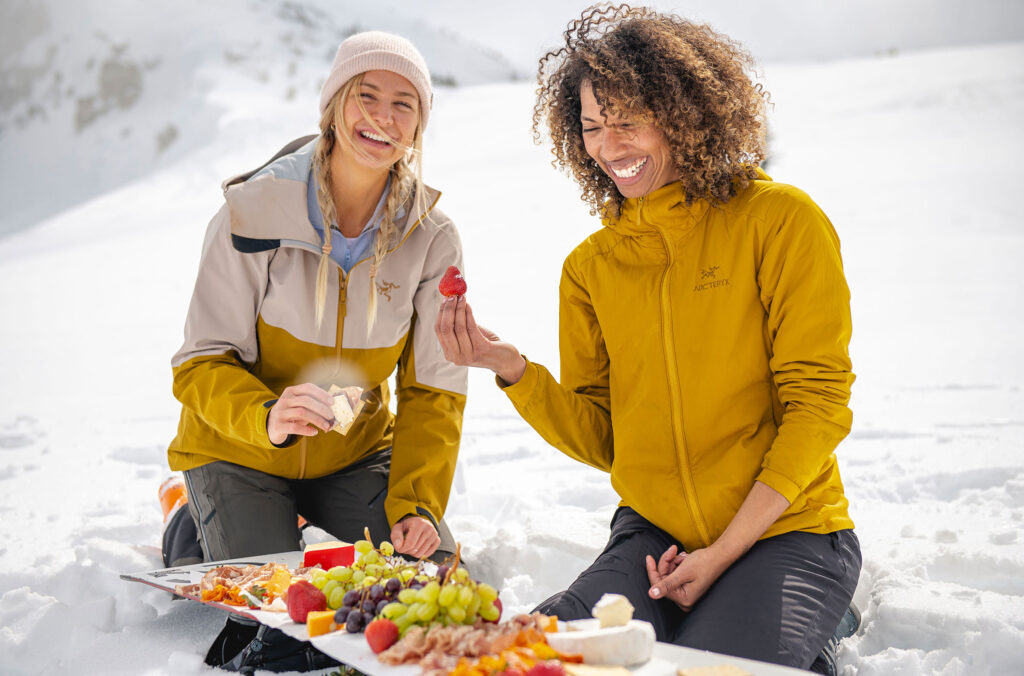 Two women eat a picnic of meats, cheese and fruits from a snowboard on Whistler Blackcomb.