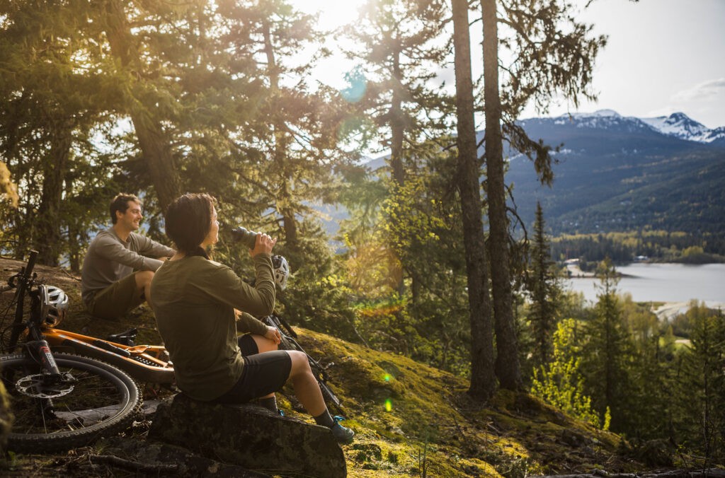 Two bike riders rest on a mossy rock looking out over the lake in Whistler.