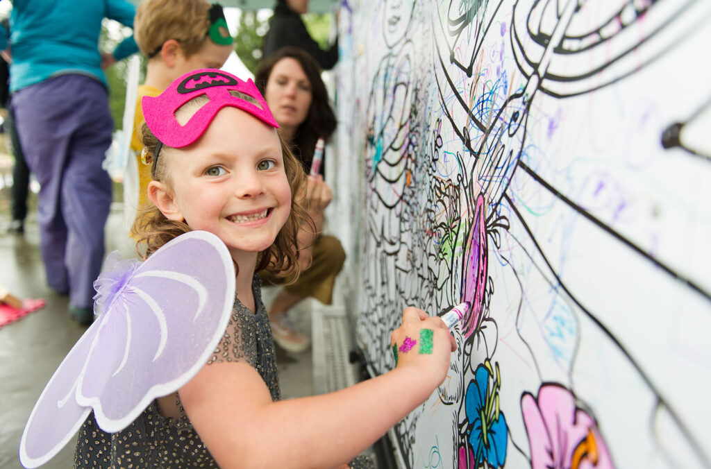 A young girl dressed as a butterfly with a batman mask draws on a large outdoor mural which is part of the Whistler Children's Festival.