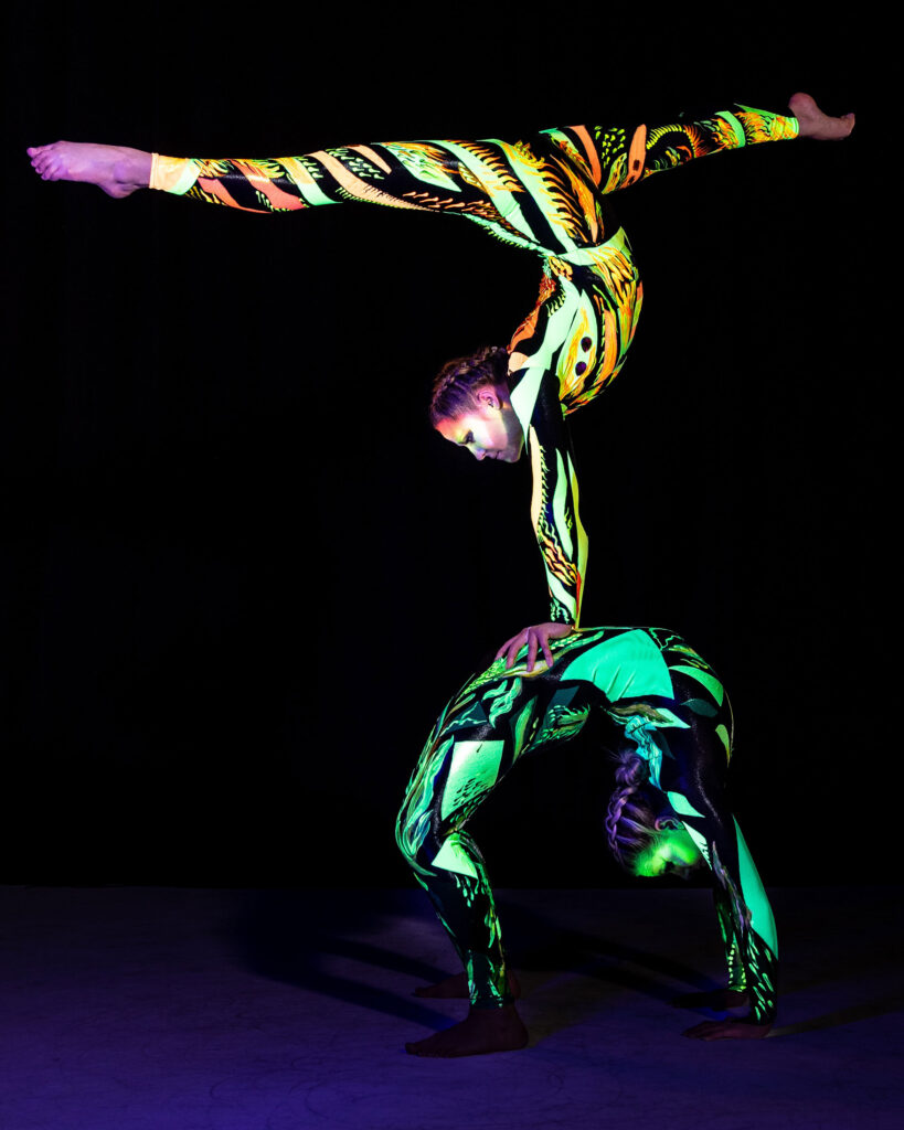 Two acrobats dressed in glowing costumes balance on one another.