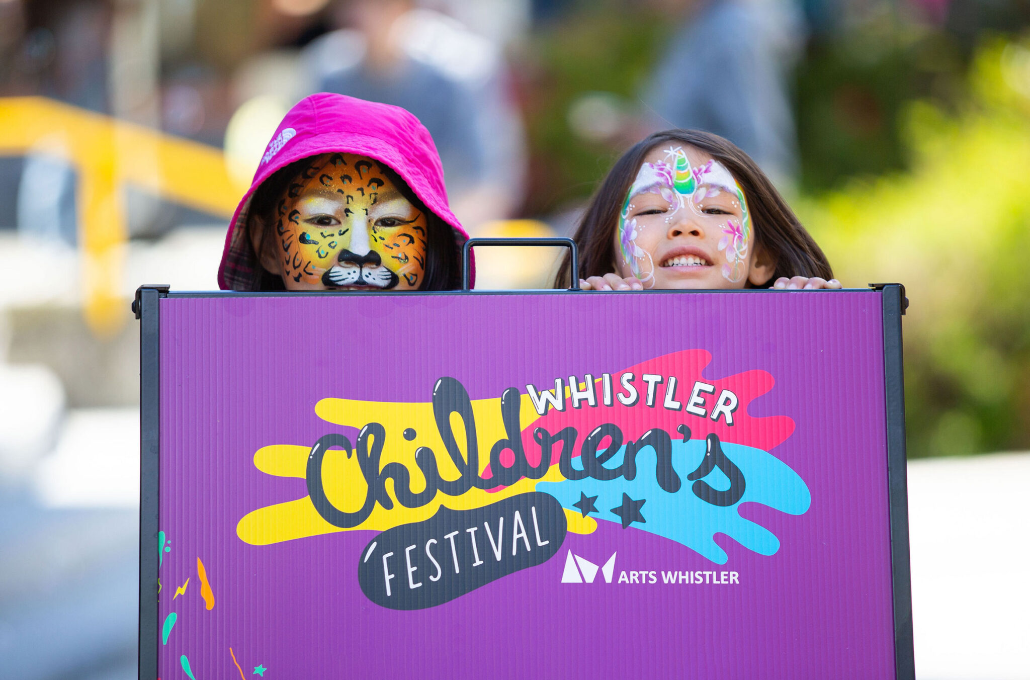 Two painted faces peek out over the top of a Whistler Children's Festival sign.