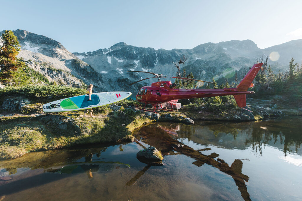 A helicopter is sat by a beautiful mountain lake as a woman gets a paddleboard out.